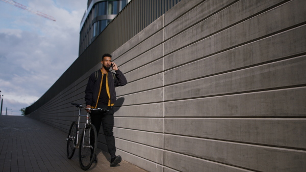 A young man commuter pushing bike and using smartphone in city.