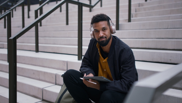 A young man sitting on bench using tablet and looking at camera in city.