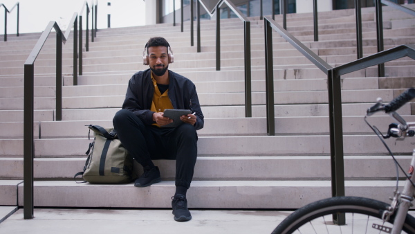 A young man sitting on bench using tablet and looking at camera in city.