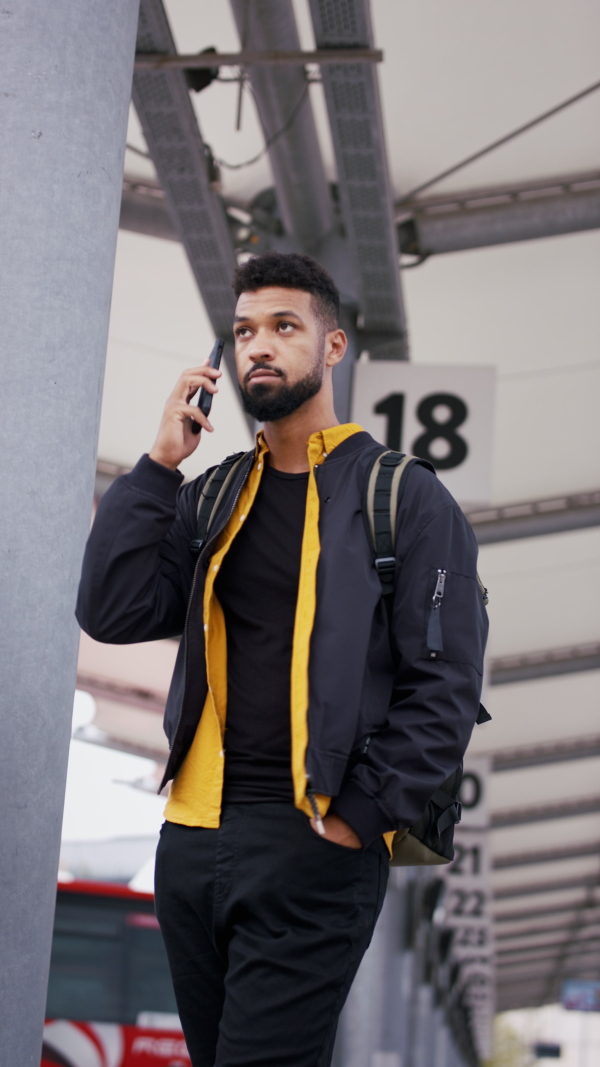 A vertical footage of young African- american man commuter waiting in bus station and using smartphone.