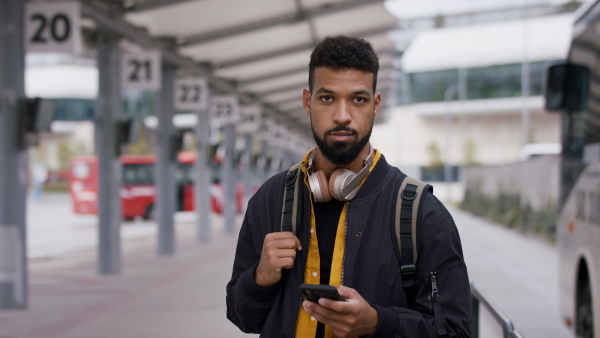 A young African- american man commuter using smartphone and looking at camera at bus station.