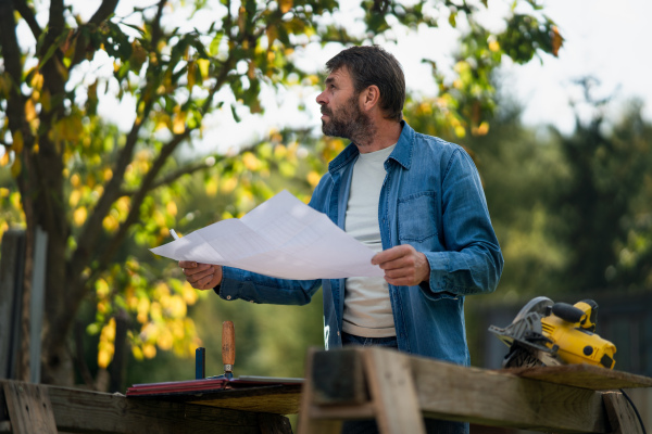 A mature man engineer with blueprints of a house outdoors.