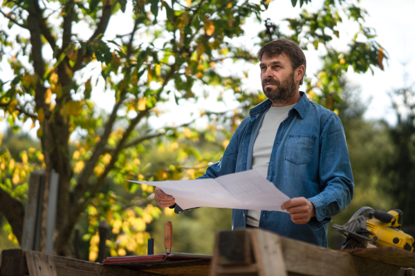 A mature man engineer with blueprints of a house outdoors.