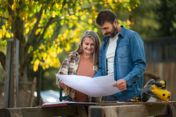 A happy mature couple with architectural blueprints of their future house, standing outdoors.