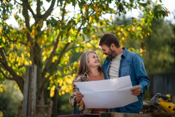 A happy mature couple with architectural blueprints of their future house, standing outdoors.