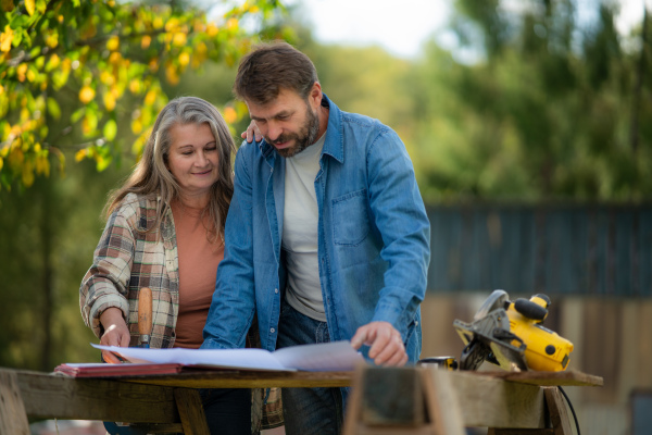 A happy mature couple with architectural blueprints of their future house, standing outdoors.