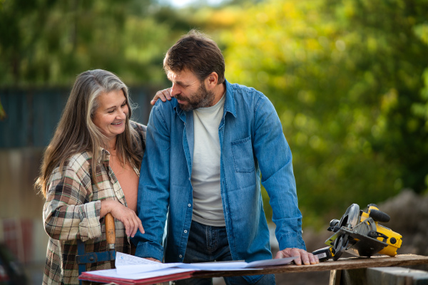 A happy mature couple with architectural blueprints of their future house, standing outdoors.