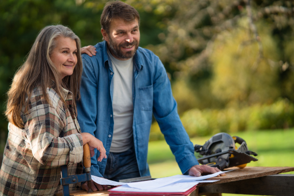 A happy mature couple with architectural blueprints of their future house, standing outdoors.