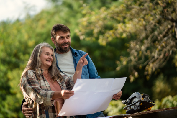 A happy mature couple with architectural blueprints of their future house, standing outdoors.