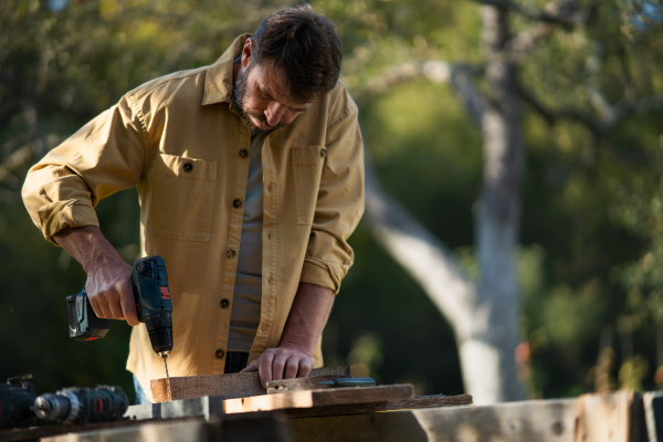 A mature handyman carpenter working in carpentry diy workshop outdoors with drill.