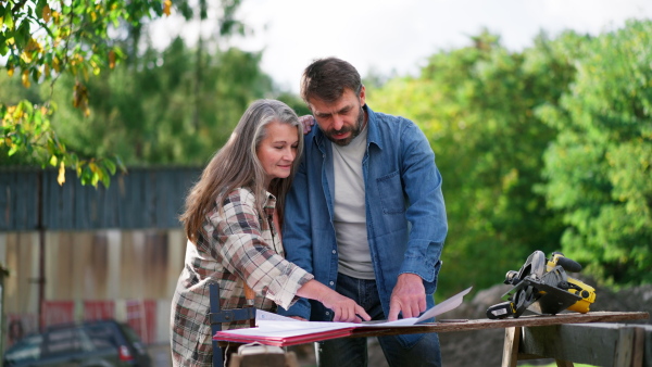 A happy mature couple with architectural blueprints of their future house, standing outdoors.