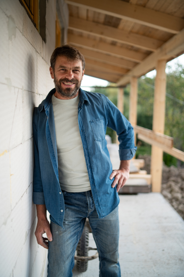 A happy mature man standing in front of his new house under construction, looking at camera.