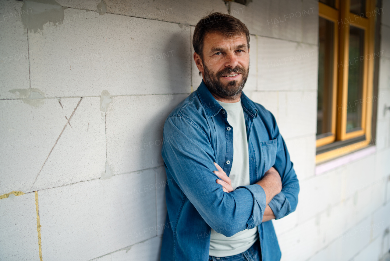 A happy mature man standing in front of his new house under construction, looking at camera.