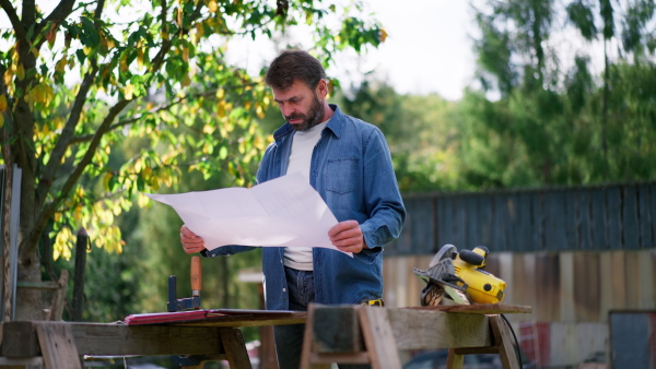 A mature man engineer with blueprints of a house outdoors.