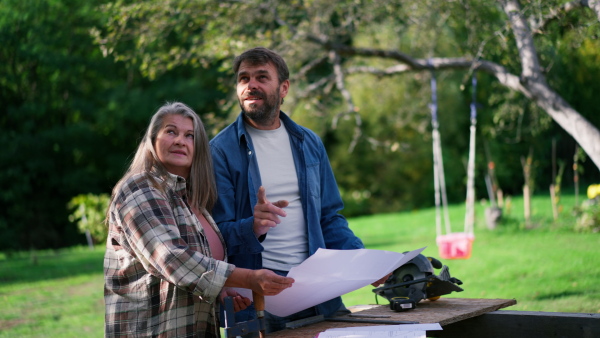 A happy mature couple with architectural blueprints of their future house, standing outdoors.