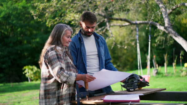 A happy mature couple with architectural blueprints of their future house, standing outdoors.