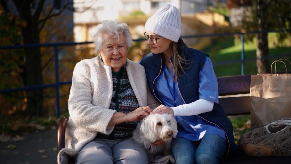 A senior woman with dog and caregiver sitting on bench outdoors in town, looking at camera.