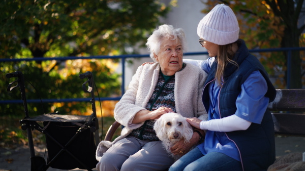 A senior woman with dog and caregiver sitting on bench outdoors in town.