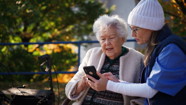 A senior woman with caregiver sitting on bench outdoors in town, using smartphone.