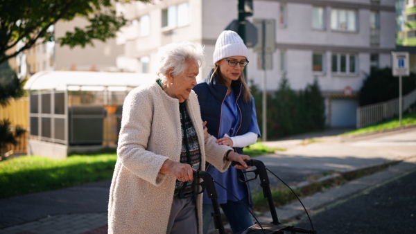 A senior woman and caregiver outdoors on a walk with walker in town, talking.