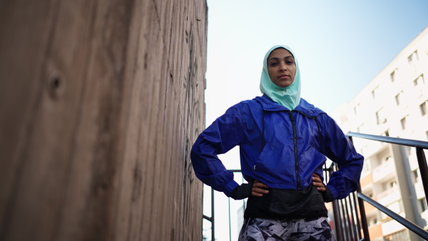 A low angle view of sporty young Muslim woman in hijab exercising outdoors in city, looking at camera.