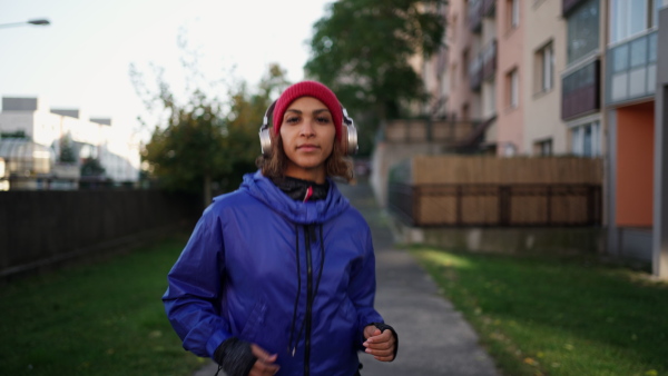 A sporty young multiracial woman with headphones running outdoors in city street, looking at camera.