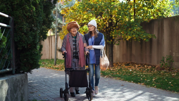 A senior woman and caregiver outdoors on a walk with walker in town, talking.