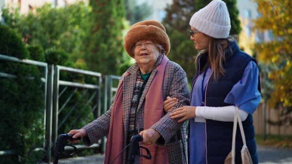 A senior woman and caregiver outdoors on a walk with walker in town, talking.