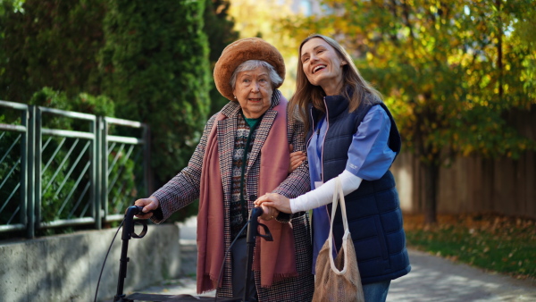 A senior woman and caregiver outdoors on a walk with walker in town, talking.