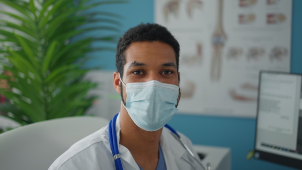 A close-up of young male doctor with face mask looking at camera in his office.