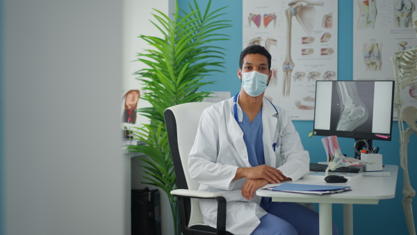 A young male doctor with face mask sitting at desk, working and looking at camera in his office.