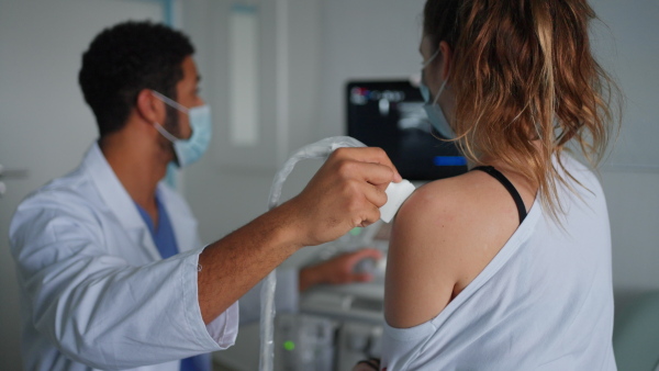 A young doctor with face mask is examining patient by using an ultrasound equipment in clinic.