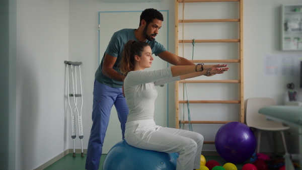 A young male physiotherapist exercising with young woman patient on ball in a physic room