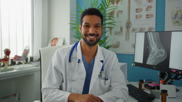 A young male doctor sitting at desk, working and looking at camera in his office.