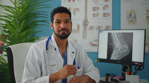 A young male doctor sitting at desk, working and looking at camera in his office.