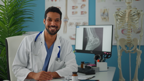 A young male doctor sitting at desk, working and looking at camera in his office.