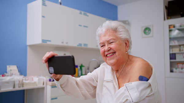 A happy senior woman with bandage on arm after vaccination taking selfie in doctor's office.