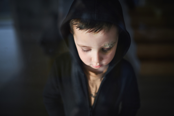 Close-up of sad small boy with cut eyebrow standing indoors at home, poverty concept. Shot through glass.