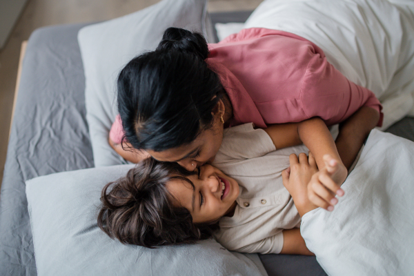 A top view of Indian mother hugging and kissin her little son lying in bed at home.