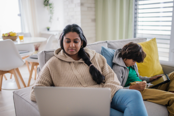 An Indian mother using laptop and her little son using tablet and sitting on sofa at home.