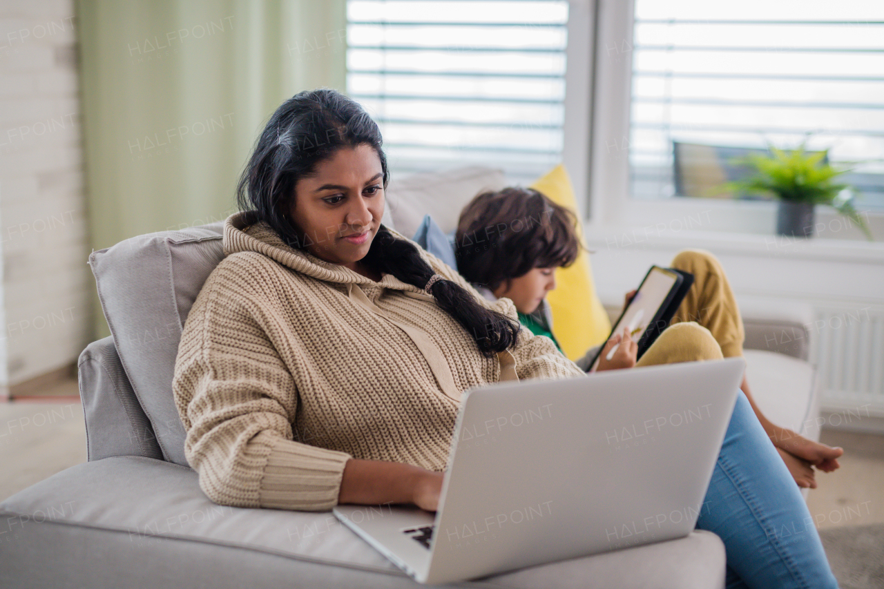 An Indian mother using laptop and her little son using tablet and sitting on sofa at home.