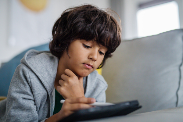 A little multiracial boy with tablet lying on sofa at home.