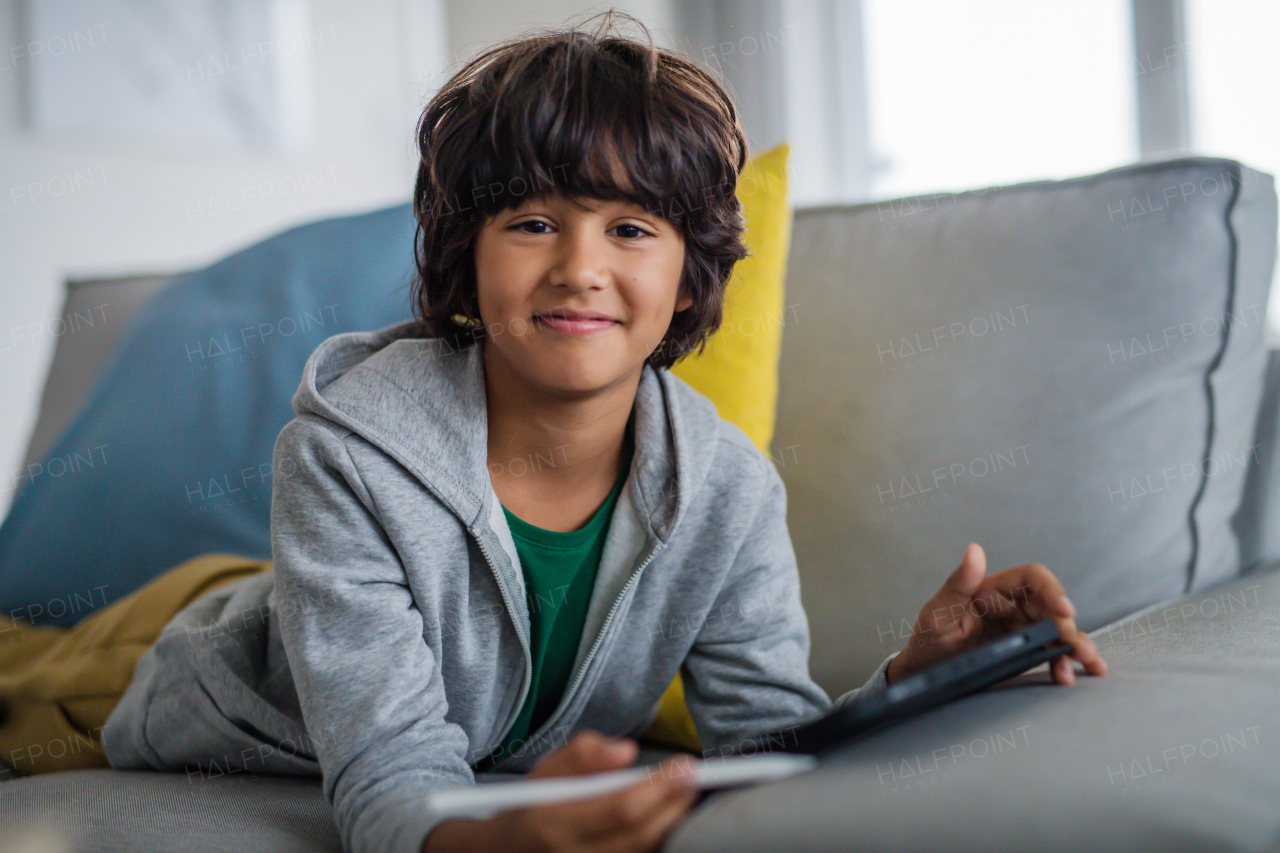 A little multiracial boy with tablet lying on sofa at home, lookingat camera.