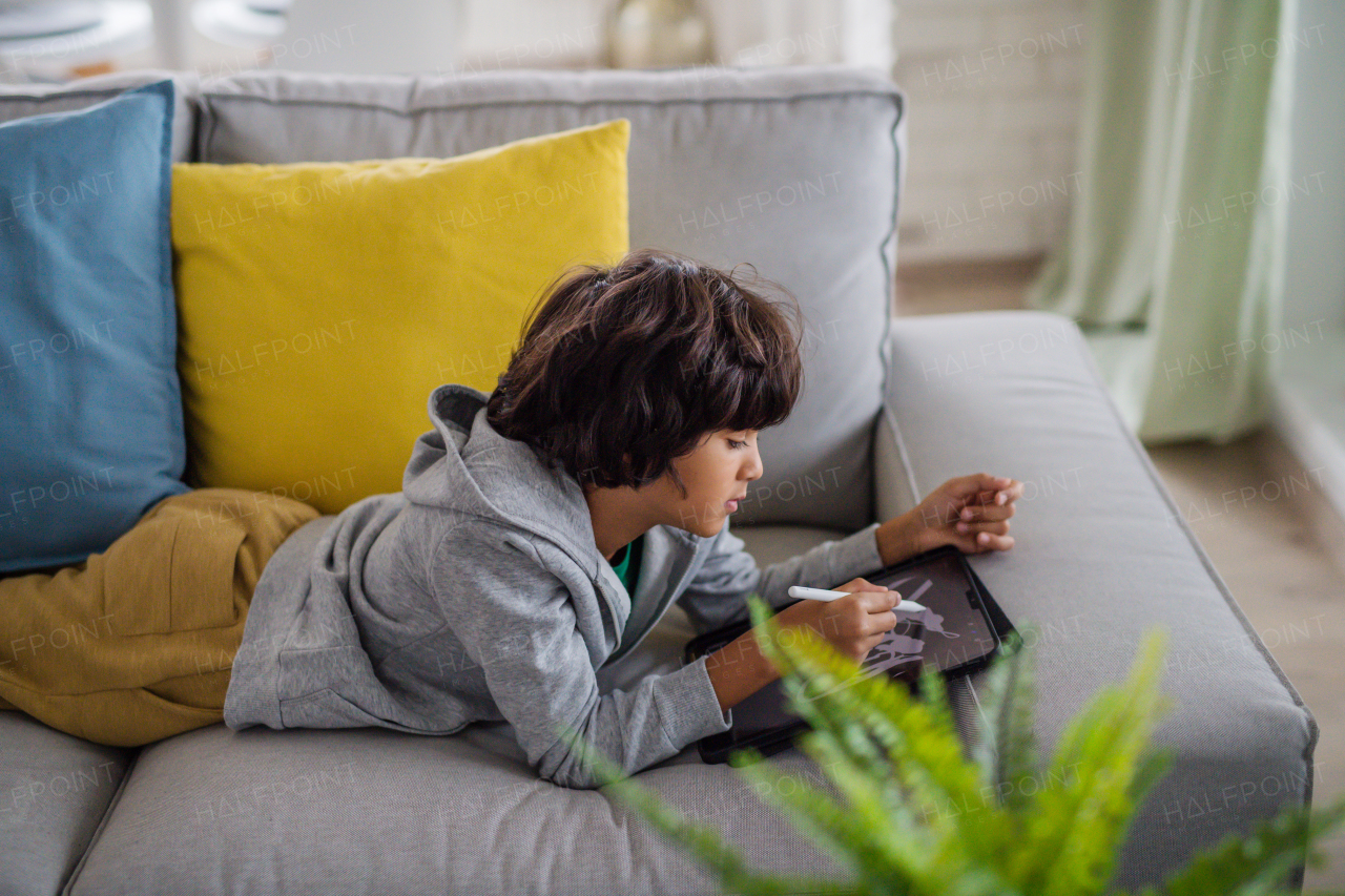 A little multiracial boy with tablet lying on sofa at home.