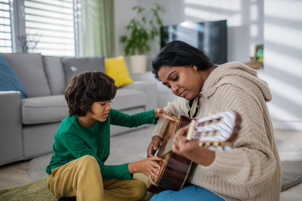 A little multiracial boy learning to play the guitar with his mother at home.