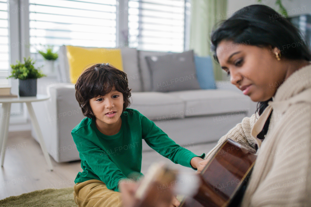 A little multiracial boy learning to play the guitar with his mother at home.