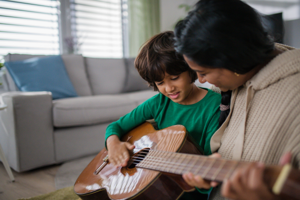 A little multiracial boy learning to play the guitar with his mother at home.