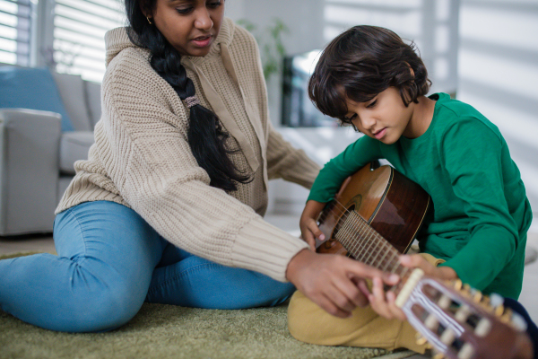 A little multiracial boy learning to play the guitar with his mother at home.