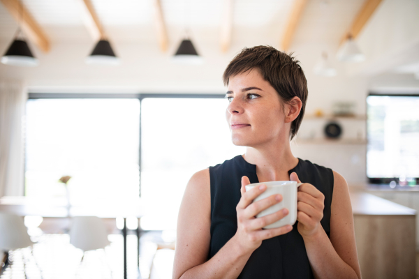 Front view of young woman with coffee standing indoors at home. Copy space.