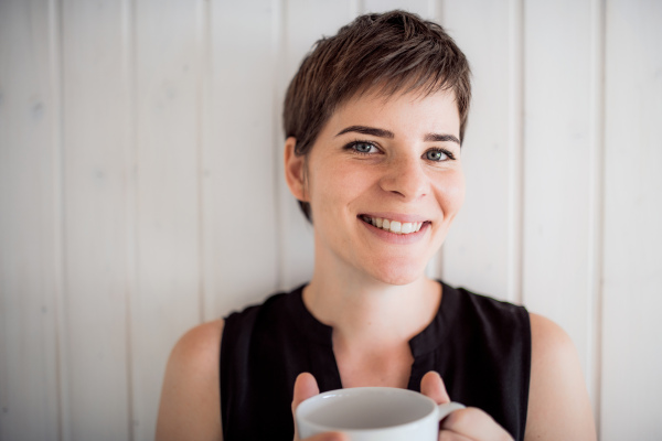 A front view of young woman with coffee standing indoors at home, leaning on wall.